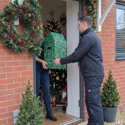 A courier carefully delivering a Tree2mydoor branded gift box to a recipient in their festively decorated home.