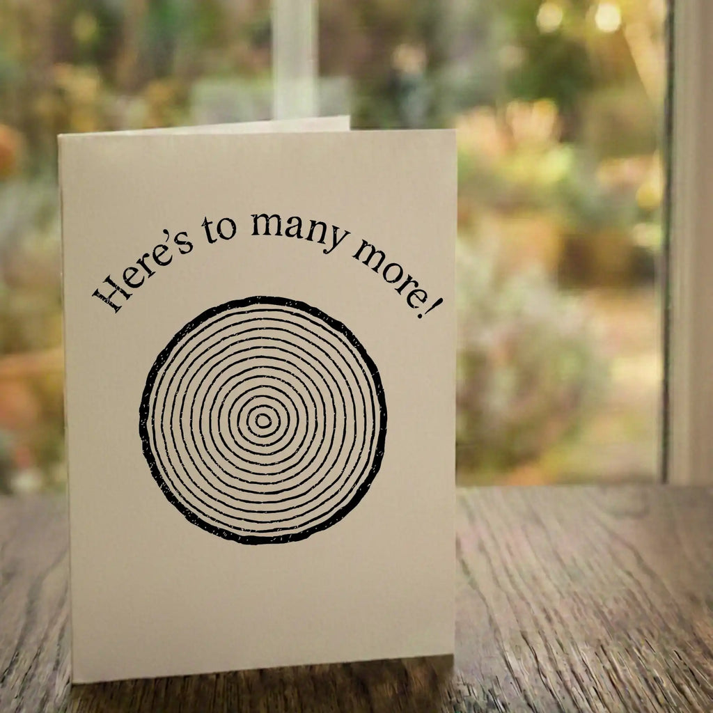 A 'Here's to many more!' card from Tree2mydoor placed on a table in front of a window with a garden view.