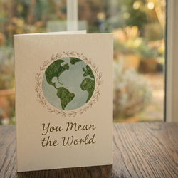 A 'You mean the world' card from Tree2mydoor placed on a table in front of a window with a garden view.